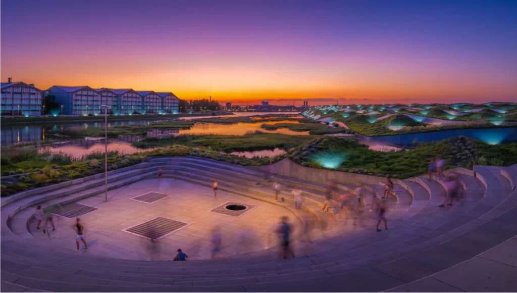 Nuvira Space architectural visualization of the Aquifer Pulse sponge city retrofit project in the Neo-London Docklands at dusk — featuring a board-formed raw concrete sunken plaza with integrated hydro-turbine grates in the foreground, intensive green-blue roofscapes on repurposed industrial warehouses in the midground, and a bioluminescent mycelium-filtered wetland waterfront edge at background, demonstrating data-driven urban resilience design, blue-green infrastructure, and post-industrial architectural transformation.