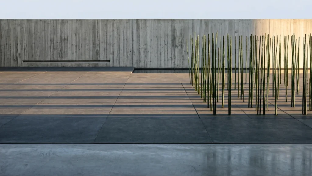 Minimalist garden architecture at golden hour featuring oversized honed anthracite limestone pavers with zero-grout flush threshold, a geometric grid of Equisetum hyemale horsetail reeds, and a mirror-flat linear water rill alongside a board-formed concrete wall — biophilic residential exterior design by Nuvira Space demonstrating monomateriality, haptic continuity, and neuro-architectural spatial clarity.