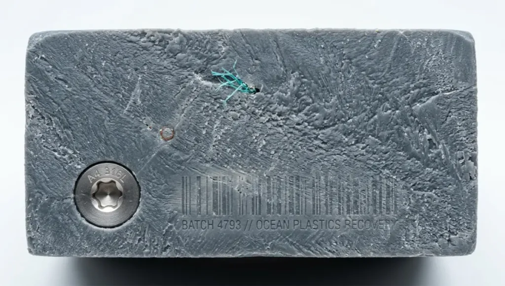Macro photography of salvaged ocean plastic lumber cross-section showing 100% ocean-recovered HDPE extrusion texture, embedded marine polymer traces including fishing net fragments, and 316L stainless steel fastener detail. Shot with 90mm macro lens at f/11 under diffused overcast lighting. Sustainable building material texture, recycled marine plastic lumber surface, ASTM-certified structural composite close-up.