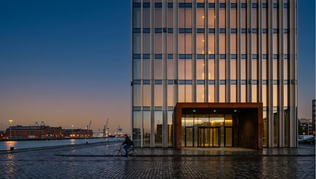 Project Meridian Node by Nuvira Space — speculative concept study of a 28-floor neuromorphic-integrated commercial tower in Rotterdam's M4H district at dusk, showing aluminum fin facade with embedded sensor nodes, cor-ten steel entrance canopy, and post-industrial waterfront context, illustrating neuromorphic sensors building management integration at architectural scale.