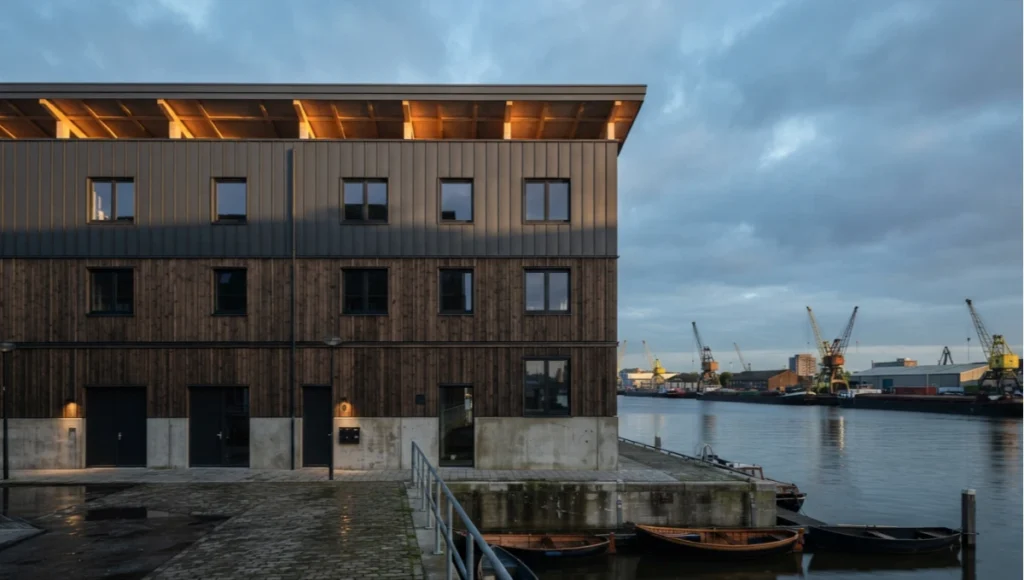Exterior architectural photo of Fjord Loom, Nuvira Space's speculative timber-framed residential concept near Rotterdam, showing charcoal cladding, exposed wood-fiber insulation board detailing, and golden-hour lighting on a regenerative infrastructure envelope.