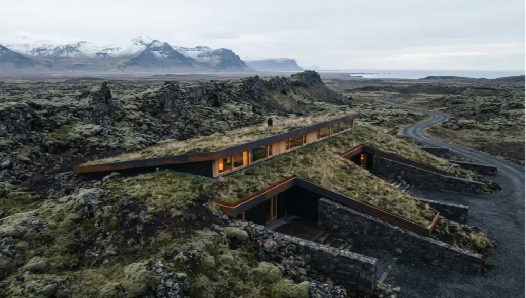 Aerial three-quarter view of Helios Refuge, a speculative Nuvira Space concept dwelling near Reykjavik, Iceland, showing its sunken profile integrated into basalt lava-rock terrain with oak-framed clerestory glazing.